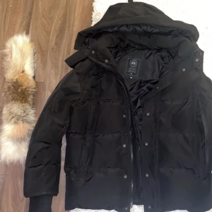 Svart dunjacka från Canada Goose - Säljer en svart dunjacka från Canada Goose med avtagbar huva och äkta pälsdetalj. Jackan har dragkedja och knappar framtill, ribbade muddar och klassisk patch på ärmen. Perfekt för kalla vinterdagar och riktigt snygg passform.