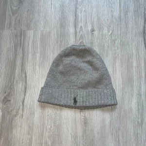 Grå mössa från Polo Ralph Lauren - Snygg grå mössa från Polo Ralph Lauren med klassisk ribbad kant och broderad svart logga framtill. Tillverkad i mjuk ullblandning som håller dig varm och stilren under kyliga dagar. Perfekt accessoar för en clean och enkel look.