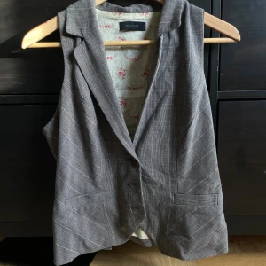 Rutig väst från Vero Moda - Stilren grå väst från Vero Moda med klassiskt rutig mönster, knappar framtill och krage. Fodret har blommigt mönster och västen har fickdetaljer samt justerbart band i ryggen. Perfekt för att lyfta din outfit med en dressad touch.