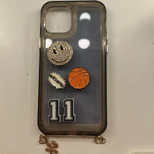 Dekorerat mobilskal för iPhone - Unikt mobilskal till iPhone med dekorativa pins: glittrande smiley, basketboll, 'AWESOME!!'-text och siffran 11. Skalets kanter är lätt tonade och det finns guldfärgade hängen längst ner. Skalet är i gott skick utan synliga repor eller skador.