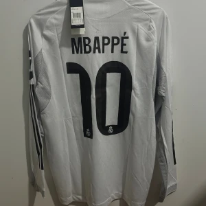 Real Madrid Mbappé långärmad tröja 25/26 Mbappe 10 Player Edition  - Säljer en officiell Real Madrid fotbollströja från Adidas med Mbappé och nummer 10 på ryggen. Tröjan är vit med svarta detaljer, gula inslag och klubbmärke på bröstet. Långärmad modell i ventilerande funktionsmaterial med HEAT.RDY-teknologi.