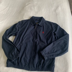 Polo Ralph Lauren jacka - Klassisk marinblå Harrington-inspirerad jacka från Polo Ralph Lauren med det ikoniska röda broderade polo-logotypen på bröstet. Tillverkad i ett lätt bomullsmaterial – perfekt för vår och höst. 	•	Storlek: S 	•	Färg: Marinblå 	•	Dragkedja framtill 	•	Resår vid ärmslut och nederkant för bra passform 	•	Två sidofickor 	•	Tillverkad i Indien 	•	Använd fåtal gånger och är i väldigt bra skick Nypris är ca 2500kr En tidlös och mångsidig jacka som passar både till vardag och mer uppklädda tillfällen.