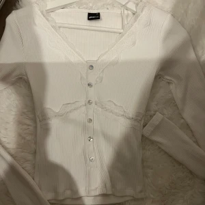 Vit ribbad topp med spets från Gina Tricot - Supersöt vit ribbad topp från Gina Tricot med långa ärmar, knappar framtill och fina spetsdetaljer vid halsringning och midja. Perfekt för dig som gillar romantisk stil och vill ha något som sticker ut lite extra.