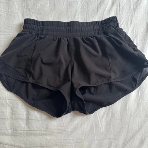 Svarta träningsshorts från Lululemon - Säljer ett par svarta träningsshorts från Lululemon med elastisk midja och innerbyxa. Shortsen har en loose passform och är tillverkade i ett lätt syntetmaterial som andas. Perfekta för löpning eller gymmet. Klassisk Lululemon-logga på sidan. Har klippt av taggen på insidan 