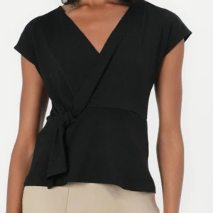 Svart omlottopp från JDY - Svart omlottopp från JDY med korta ärmar och snygg knytning i sidan. Toppen har v-ringning och en stilren, enkel design som passar perfekt till jeans eller kjol. 