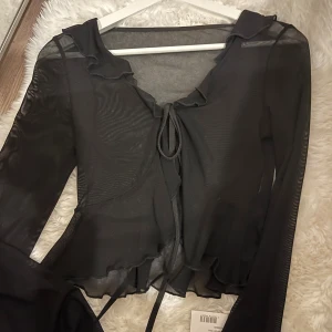 Svart mesh blus med volang och knyt - Säljer en svart transparent meshblus med långa ärmar, volangdetaljer längs kanterna och knyt framtill. Blusen har en croppad passform och är perfekt för att skapa en edgy och trendig look.