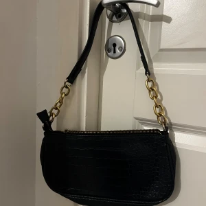 Mörkblå axelväska med kedjedetaljer - Snygg Mörkblå axelväska med krokopräglat mönster och guldfärgade kedjor på axelremmen. Väskan har dragkedja upptill och ett innerfack med extra dragkedja. Perfekt storlek för det viktigaste och ger en cool vibe till din outfit. Nästan helt oanvänd, vid snabb affär kan jag gå ner i pris