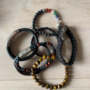 Mix av 6 coola armband i olika stilar - Säljer ett set med 6 armband: flätade svarta läderarmband med metallspänne, pärlarmband med tigeröga och träpärlor, samt ett färgglatt pärlarmband med buddha-detalj. Fråga gärna efter mått är inte säker på storlekar