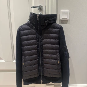 Moncler cardigan - Säljer en mörkblå cardiganfrån Moncler med stickade ärmar och huva. Jackan har quiltad framsida, dragkedja och praktiska fickor med knappstängning. Snyggt Moncler-märke på ärmen och cool urban look. Perfekt för dig som gillar exklusiva streetwear-vibbar.