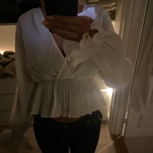 Vit omlott blus med volangärm - Säljer en vit omlott blus med v-ringning och långa volangärmar. Blusen har ett plisserat tyg med volangkant nedtill. Superfin och går även att vika upp ärmarna för en annan stil. 💕
