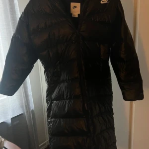 Svart lång dunjacka från Nike - Lång svart dunjacka från Nike med huva och dragkedja hela vägen. Perfekt för kalla dagar när du vill hålla dig varm och stilsäker. Nyspris: 3395kr. HELT oanvänd med prislapp kvar.