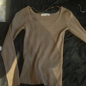 Beige ribbad långärmad topp Samsa - Säljer en beige ribbad långärmad topp från Samsa Collection. Toppen har en snygg, något fyrkantig ringning och är figurnära i modellen. Perfekt att styla med jeans eller kjol för en clean och trendig look.