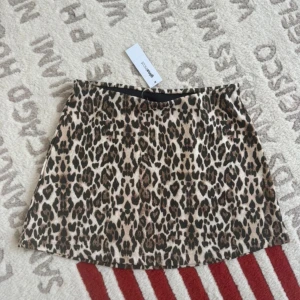 Leopardmönstrad kortkjol från Gina Tricot  - Snygg kortkjol från Gina Tricot med leopardmönster i beige och bruna toner. Kjolen har en rak passform och är perfekt för dig som vill sticka ut med ett djuriskt print. Materialet är mjukt och följsamt, vilket gör den bekväm att bära hela dagen.