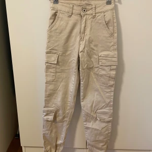 Beige cargopants med fickor - Säljer ett par beige cargopants med flera praktiska fickor på benen och bak. Byxorna har elastiska benslut och klassisk midja. 