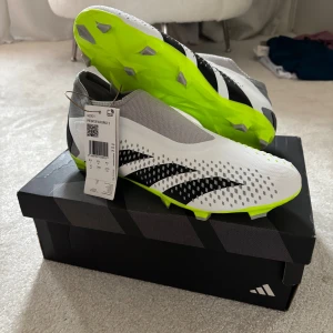 Adidas Predator Accuracy 3 fotbollsskor Storlek 44 - Adidas Predator Accuracy 3 fotbollsskor i vit, grå och neon-gul färg med svarta detaljer. Skorna har en strömlinjeformad siluett, texturerad ovandel och platt sula med dobbar för gräsplan. Materialet är syntet och mesh för bra ventilation och passform.