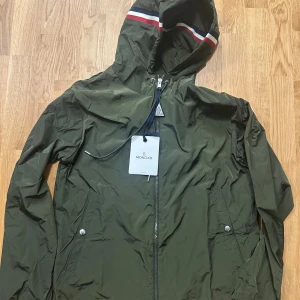 Grön vindjacka från Moncler med huva - Snygg grön vindjacka från Moncler med huva och dragkedja framtill. Jackan har Moncler-logga på ärmen och coola ränder i rött, vitt och blått på huvan. Lätt material i polyester, perfekt för blåsiga dagar. Fickor med knappstängning och justerbar snörning vid huvan.