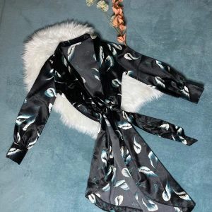 Svart omlottklänning med blommönster - Snygg svart omlottklänning med fjäderliknande mönster i vitt och blått. Klänningen har lång ärm, breda manschetter och ett knytband i midjan som ger en cool siluett. Materialet är glansigt och känns lyxigt, perfekt för dig som gillar att sticka ut.