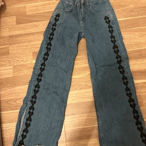 Wide jeans från Noon med broderi - Säljer ett par blå jeans från Noon i wide fit med coolt svart broderi längs benen. Klassisk femficksmodell med hög midja och en liten röd lönn på bakfickan. Jeansen är tillverkade i jeansmaterial och har en snygg, avslappnad look.