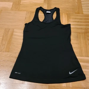 Svart Nike Pro Dri-Fit linnetopp - Svart linnetopp från Nike Pro med Dri-Fit-teknologi, perfekt för träning. Modellen är ärmlös med brottarrygg och har ventilerande meshpaneler för extra komfort. Snyggt silverfärgat Nike-logga framtill. Sitter tight och följer kroppen.