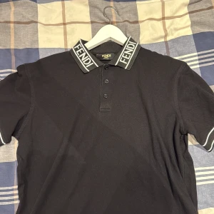 Svart Fendi piké med logga på kragen - Svart pikétröja från Fendi med tydlig logga på kragen och vita ränder på ärmslut och krage. Klassisk passform, tre knappar framtill och tillverkad i bomull. Perfekt för dig som vill ha en stilren och lyxig look.