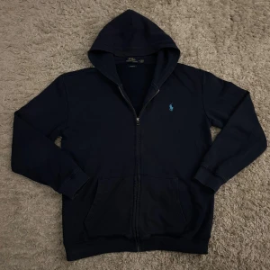 Marinblå hoodie från Polo Ralph Lauren - Snygg marinblå hoodie från Polo Ralph Lauren i bomull med dragkedja framtill och huva. Klassisk broderad logga i ljusblått på bröstet och två fickor. Custom fit-modell med långa ärmar och ribbade muddar. Perfekt för chill dagar.