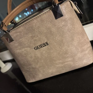 Beige handväska från Guess - Snygg beige handväska från Guess med stilrena silverdetaljer och robusta handtag i ljusbrunt. Fick i present men aldrig använts. 