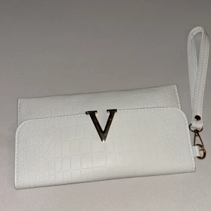 Vit kuvertväska med V-detalj - Snygg vit kuvertväska med krokodilpräglad yta och guldfärgad V-detalj på framsidan. Väskan har ett avtagbart handledsband med guldfärgad hake. Perfekt storlek för det viktigaste och stilren design som sticker ut.