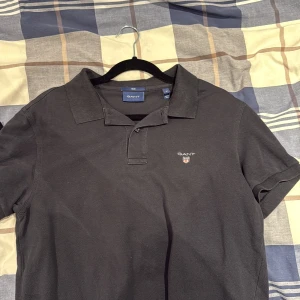 Svart pikétröja från GANT, slim fit - Svart pikétröja från GANT i slim fit med klassisk krage och korta ärmar. Tröjan har GANT-logga broderad på bröstet och är tillverkad i mjuk bomull som känns skön mot huden. Perfekt för dig som gillar stilrena och enkla plagg.