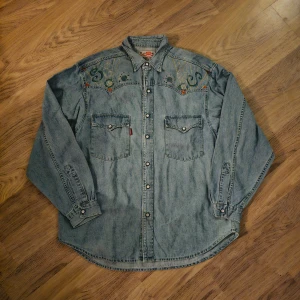 Broderad jeansskjorta från Crocker - Säljer en ljusblå jeansskjorta från Crocker med cool broderad design och glitterdetaljer på axlarna. Skjortan har två bröstfickor med knappar och klassisk krage. 