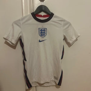 England Nike fotbollströja vit - Snygg England fotbollströja från Nike i vit polyester med blått och rött på kragen och sidorna. Tröjan har korta ärmar, broderat England-emblem med tre lejon och en silverfärgad stjärna ovanför. Perfekt för dig som vill visa support för landslaget.