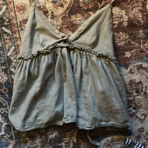 Grå linnetopp med volang från Zara - Säljer en grå linnetopp från Zara i storlek M (men jag skulle säga att den är xs)Toppen har smala axelband, v-ringad front och volangdetaljer längs bröstet. Perfekt för dig som gillar chill och stilren vibe.