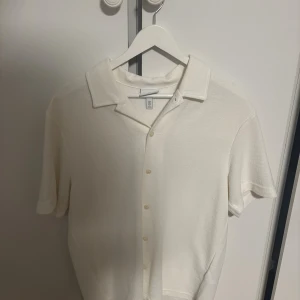 Vit kortärmad skjorta med knappar från H&M - Vit kortärmad skjorta från H&M med klassisk krage och knäppning framtill. Skjortan har en avslappnad passform och är tillverkad i ett mjukt bomullstyg som känns skönt mot huden. Perfekt för en clean och stilren look.