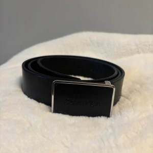 Svart skärp från Calvin Klein - Snyggt svart bälte från Calvin Klein med stilren, rektangulär metallspänne där loggan är ingraverad. Bältet är gjort i slätt skinn och har en klassisk, minimalistisk design som passar till jeans eller kostymbyxor. Perfekt för dig som gillar cleana accessoarer.