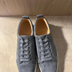 Grå mocka sneakers Christian Louboutin - Snygga grå sneakers från Christian Louboutin i mjuk mocka med klassisk rund tå och snörning. Skorna har den ikoniska röda sulan med Louboutin-logga och vit kant. Perfekt för dig som gillar exklusiva detaljer och stilren design.