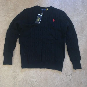 Ralph Lauren - Svart stickad tröja från Polo Ralph Lauren med klassisk kabelstickning och röd broderad logga på bröstet. Rund hals och långa ärmar, perfekt för dig som gillar stilrena och tidlösa plagg. Snygg att matcha med jeans eller chinos.