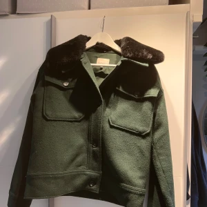 Grön overshirt med pälsig krage från Noella - Snygg mörkgrön overshirt från Noella med stor svart pälsig krage. Jackan har två stora bröstfickor med knappar, rak passform och långa ärmar. Materialet är ullmix och detaljerna ger en cool och trendig vibe. Perfekt för dig som gillar statement-plagg.