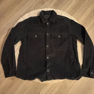 Svart manchester overshirt jackliknande - Säljer en svart overshirt i manchester med klassisk krage, två stora bröstfickor med lock och knappar framtill. Skjortan har långa ärmar och en loose passform. Den är i tjockare material så kan med fördel användas som jacka 