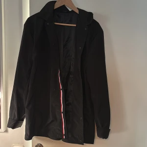 Svart regnjacka från YI&CONCEPT - Svart regnjacka med huva från YI&CONCEPT. Jackan har långa ärmar, knäppning framtill och en insida med svart foder. Snygg detalj med rödvit rand längs insidan av knäppningen. Perfekt för blöta dagar.