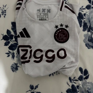 Vit Ajax fotbollströja Adidas S - Ajax fotbollströja i vit med mörkröda detaljer och geometriskt mönster. Klassisk rund hals, korta ärmar och broderat klubbmärke på bröstet. Adidas-logga och Ziggo sponsortryck framtill. Tillverkad i lätt och ventilerande polyester.