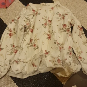 Blommig långärmad tröja från Newbie - Supersöt långärmad tröja från Newbie i storlek 110/116. Tröjan är cremevit med romantiskt blommigt mönster i rosa och grönt. Den har volangdetaljer nertill och är tillverkad i mjuk bomull, perfekt för vår och sommar.
