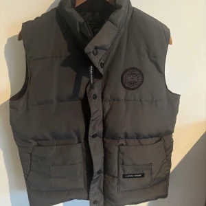 Grå dunväst från Canada Goose M  - Snygg grå dunväst från Canada Goose med hög krage, två stora fickor framtill och diskret svart logotyp på bröstet. Västen har knappar och dragkedja framtill och är perfekt för lager-på-lager-stil under kyliga dagar.