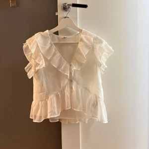 Vit blus med volanger och knappar från Nelly - Supersöt vit blus med volanger längs axlar och nederkant, samt v-ringning och knappar framtill. Blusen är kortare i modellen. 