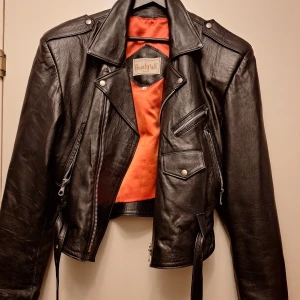 Svart skinnjacka från Beautiful - Säljer en svart skinnjacka från Beautiful med klassisk bikerstil. Jackan har flera dragkedjor, snedställd front, axelklaffar och bälte i midjan. Insidan är fodrad i orange tyg. Perfekt för dig som vill ha en edgy och tidlös look.