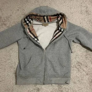 Grå Burberry hoodie med dragkedja - Snygg grå hoodie från Burberry med klassiskt rutigt foder i huvan. Tröjan har dragkedja framtill, snörning vid huvan och två fickor. Långärmad och med broderad Burberry-logga på bröstet. Perfekt för dig som gillar stilrena och lyxiga streetwear-plagg. Äkta fick tidig jul present vi ska resa utomlands 