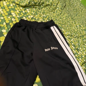 Palm Angels svarta mjukisbyxor - Svarta mjukisbyxor från Palm Angels med vita sidoränder och broderad logga på benet. Byxorna har resår i midjan och en avslappnad passform, perfekta för en streetwear-look.
