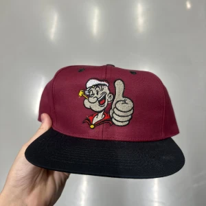 Vinröd keps med Popeye brodyr - Snygg vinröd keps med svart skärm och grön undersida. Framtill finns en broderad bild på Popeye som ger tummen upp, med tydliga färger och detaljer. Kepsen har klassisk snapback-modell och är tillverkad i slitstark polyester.