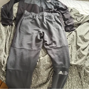 Grå träningsbyxor från The North Face - Säljer ett par grå träningsbyxor från The North Face med diskret mönstrad yta och elastisk midja. Byxorna har logga på benet och är tillverkade i ett syntetmaterial som andas. Perfekta för aktiva dagar och sport.