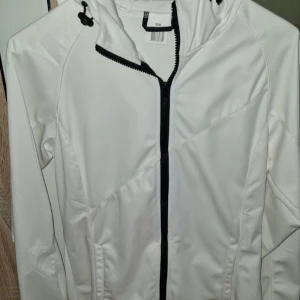 Vit vindjacka med huva S/M - Fräsch vit vindjacka i 100% polyester med svart dragkedja och justerbar huva. Jackan har lång ärm och stilrena linjer, perfekt för dig som gillar enkel och clean look. Finns fickor på sidorna och snygga detaljer vid huvan.