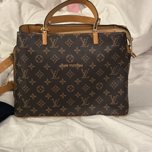  Monogram handväska - Snygg handväska med klassiskt monogrammönster i brunt och beige. Väskan har både handtag och axelrem, guldfärgade detaljer och dragkedja upptill. Perfekt storlek för att få plats med allt du behöver.