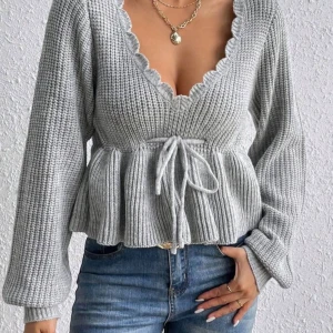 Grå stickad tröja med volangkant - Superfin grå stickad kofta med volangkant längs v-ringningen och knyt i midjan. Koftan har långa ärmar och en peplum-liknande form som ger en söt och trendig look. Perfekt att styla med jeans eller kjol för en chill vibe.